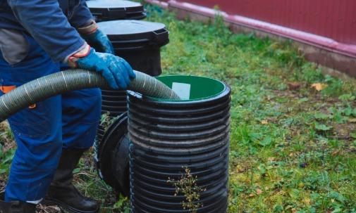 A worker wearing blue clothing and gloves holding a wide hose inside a black septic tank opening on grass.