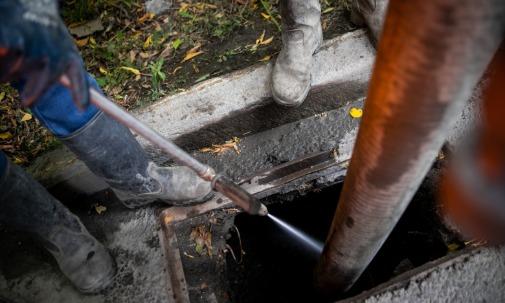 A water jet cleaning debris inside a drain opening near gray boots, a large hose, concrete curb, and leaves.