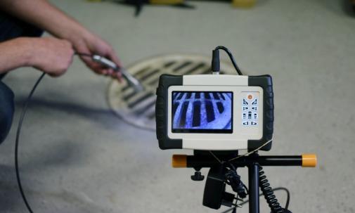 A person's hands guiding a cable near a floor drain while an inspection monitor displays drain grate bars.