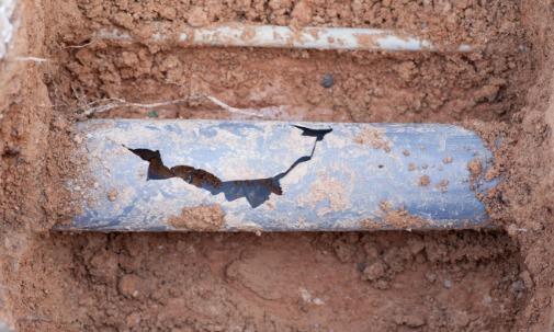 A cracked blue pipe lying in a dirt trench with loose soil and small roots around the damaged opening.