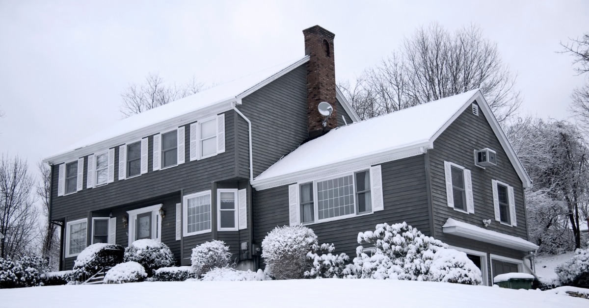 A gray two-story house with white shutters, snow-covered shrubs, a chimney, and a snowy front yard and bare trees.