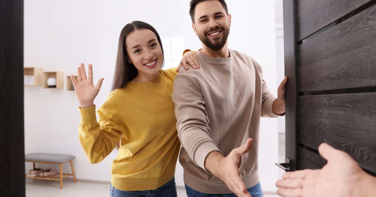 A smiling woman waving beside a smiling man reaching for a handshake near an open front door in a bright entryway.
