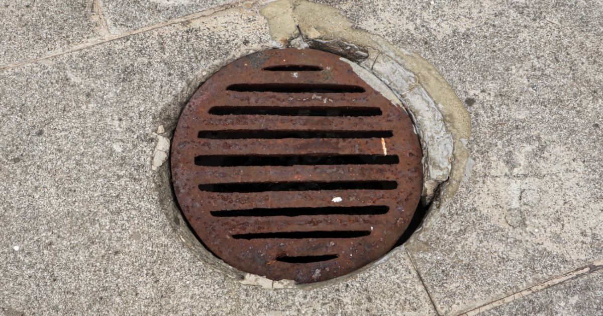 A rusty round metal drain cover with long slots embedded in worn concrete pavement near broken edges.